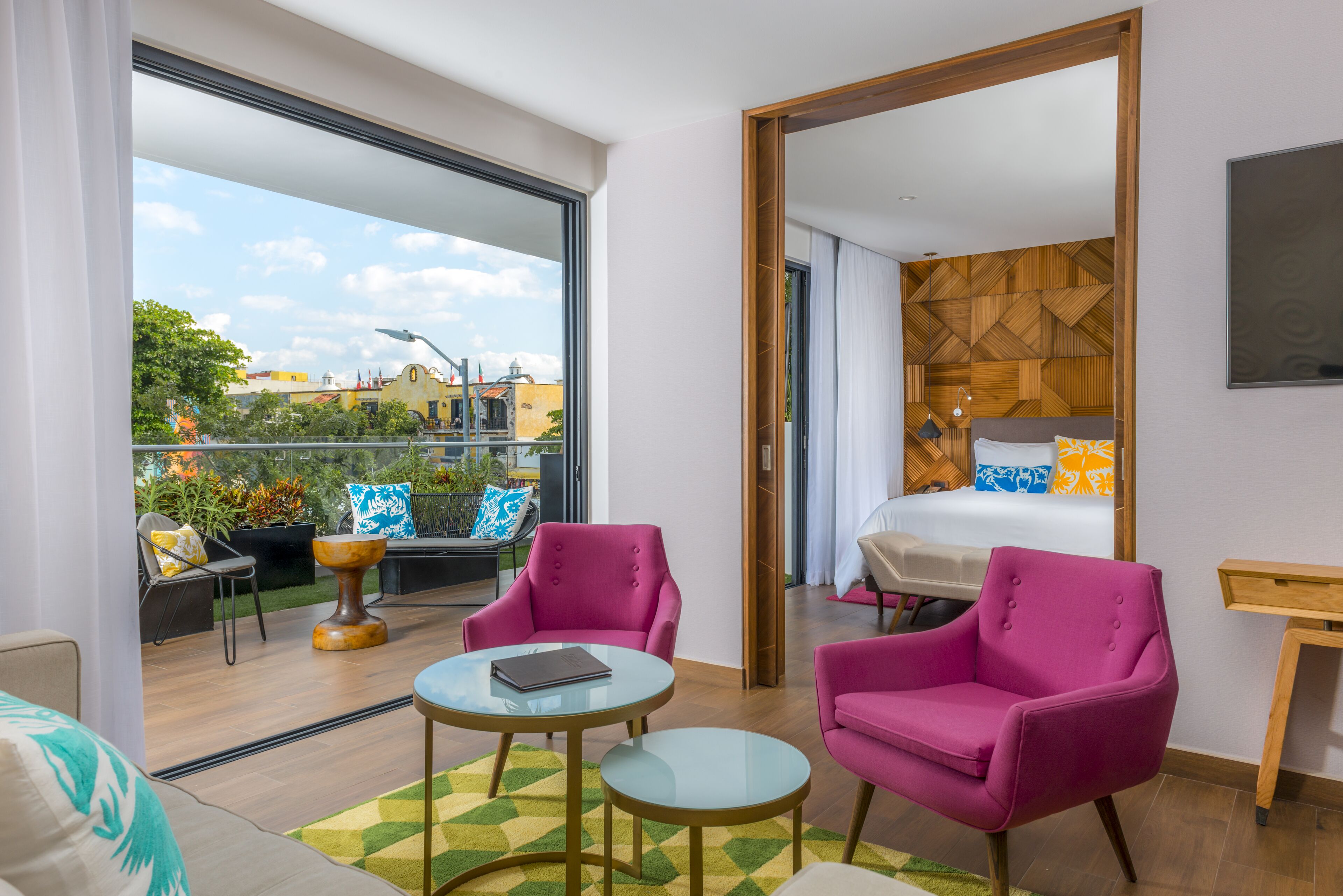 Una habitación de hotel moderna con balcón, una cama con almohadones estampados, una mesita redonda pequeña y dos sillones rosados.