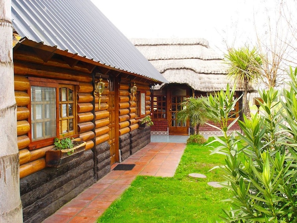 Eine Blockhütte mit einem Strohdach, Holzfassade und einem kleinen Garten mit Pflanzen und einem Weg.