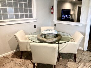 A dining area with a glass table surrounded by four white chairs, a fire extinguisher on the wall, and a television mounted above the table. - EAST MONTGOMERY GETAWAY KING BEDS COVERED PATIO SMART TVS KITCHEN WELL STOCKED (Montgomery)