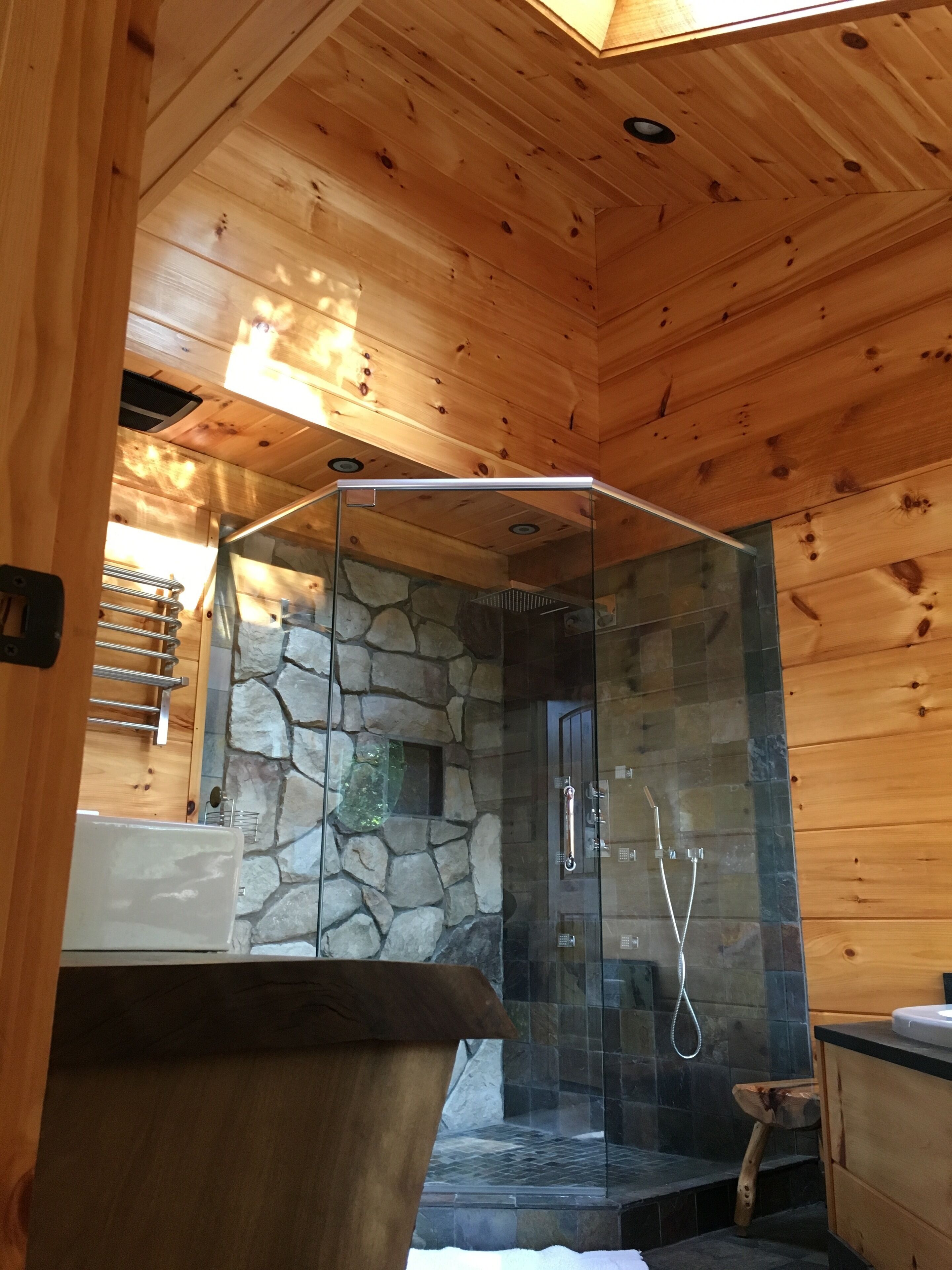 Baño de cabaña de madera con cabina de ducha de vidrio, pared de piedra y muebles de madera.