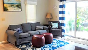 Una sala de estar con un sofá gris, una mesita y una silla. Hay una ventana con persianas, una lámpara y un cuadro en la pared.