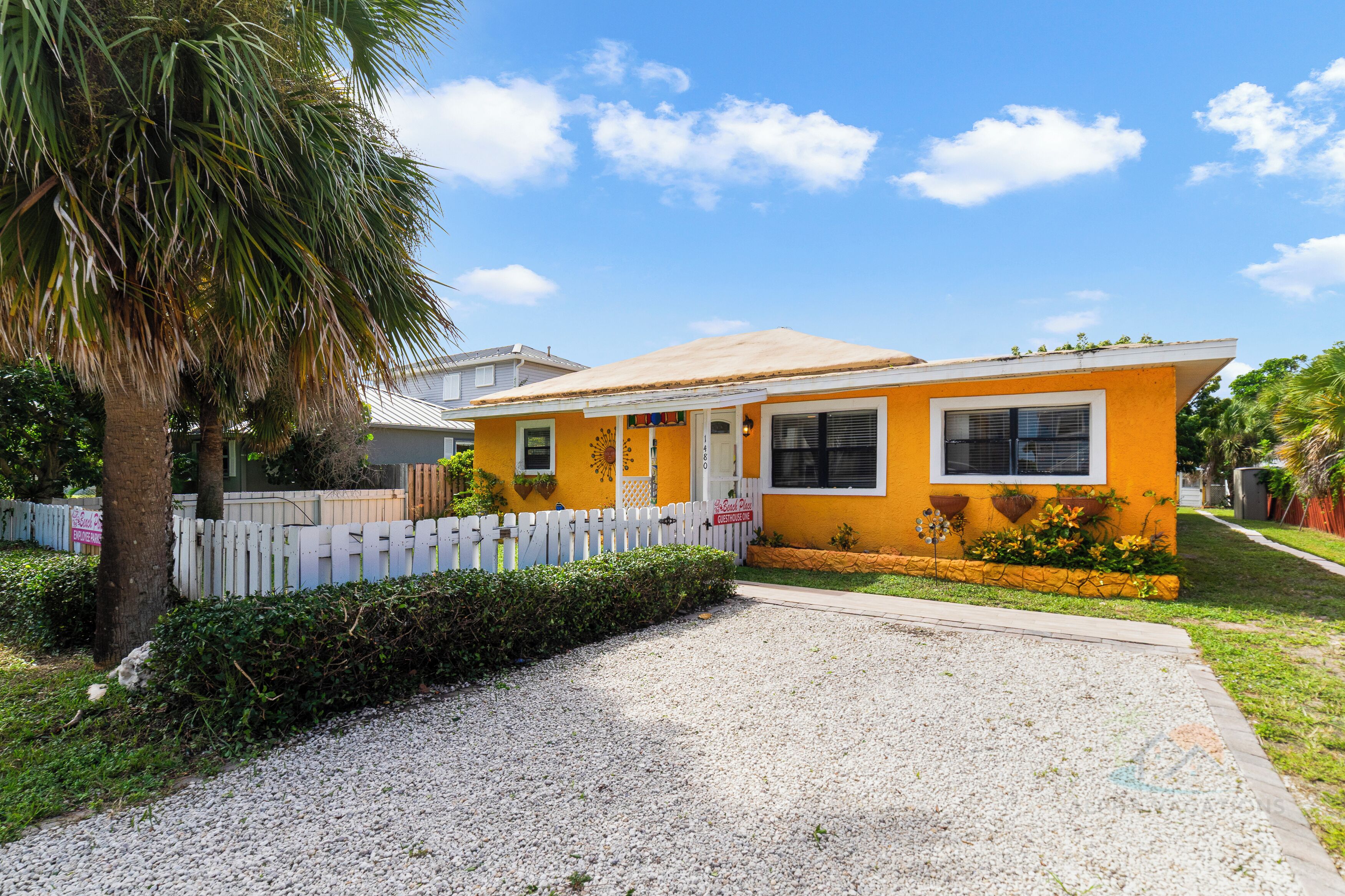 A yellow house with a white picket fence, a gravel driveway, and a palm tree.