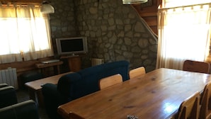 A wooden dining table with chairs, a blue sofa, a television, and a stone wall. - Cabañas La Pampita (Villa La Angostura)