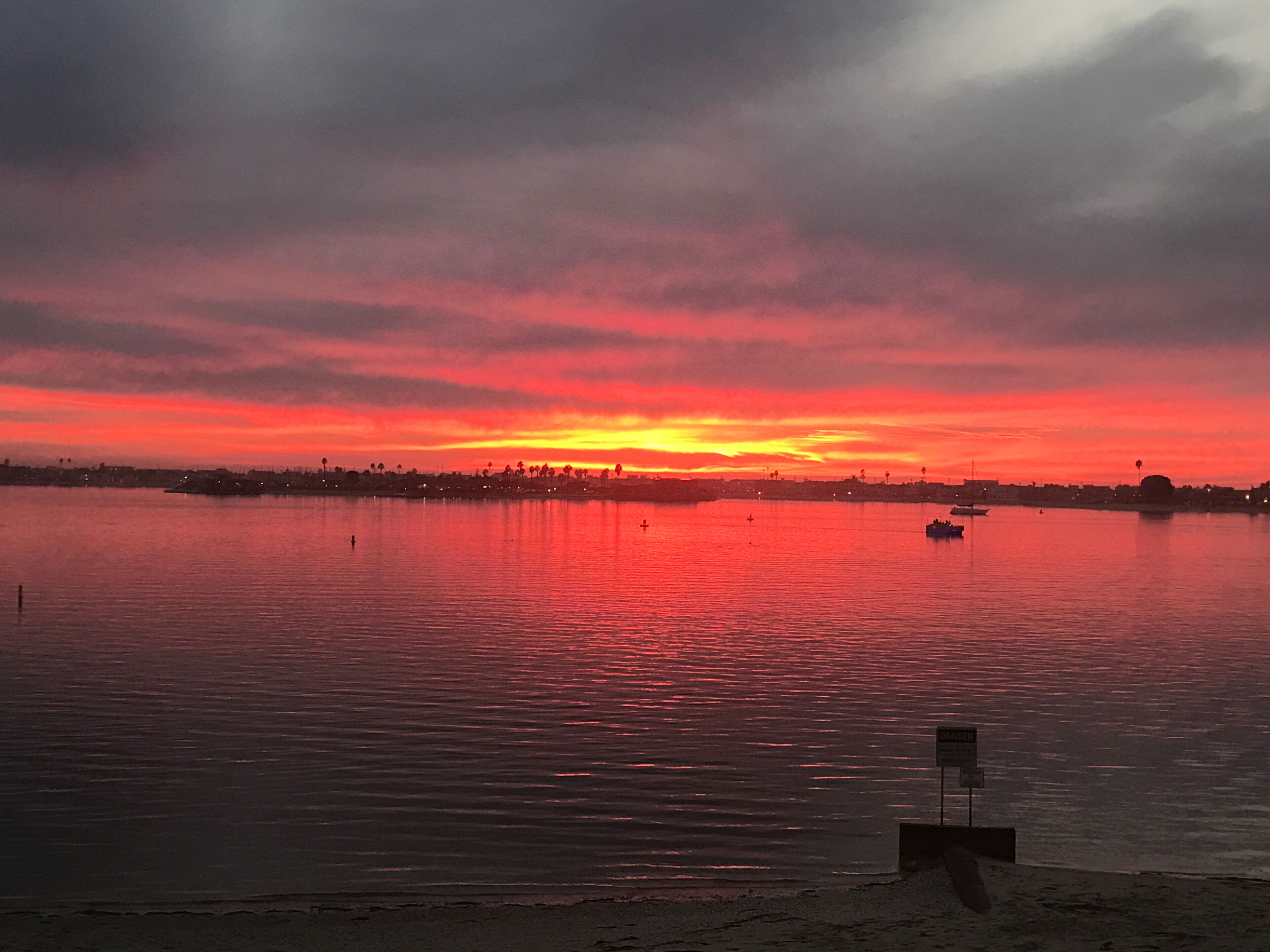 Ein Sonnenuntergang über ruhigem Wasser, im Hintergrund eine Küstenlinie und ein Schild, das auf einen Gefahrenbereich hinweist.
