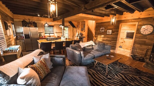 Ein gemütliches Blockhaus-Interieur mit Wohnbereich, der eine Sitzecke und einen Couchtisch sowie eine Küche mit Kühlschrank und Esstisch und einer Holzwand mit einer Uhr zeigt.