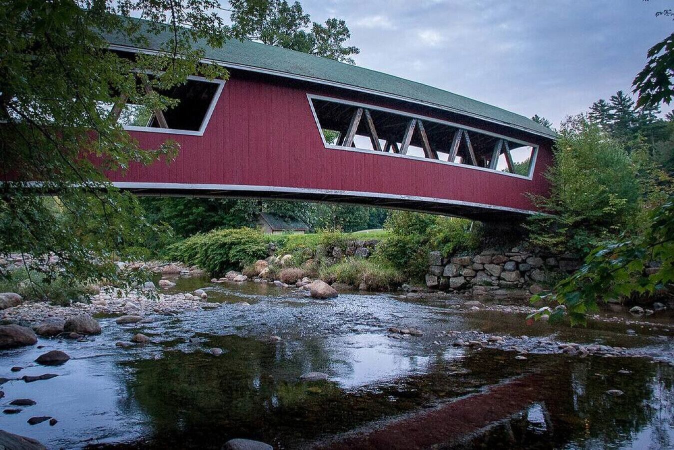 A covered bridge with a stone foundation, a stream below, and surrounding greenery.