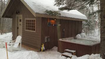 A wooden cabin with a hot tub and two Adirondack chairs outside.