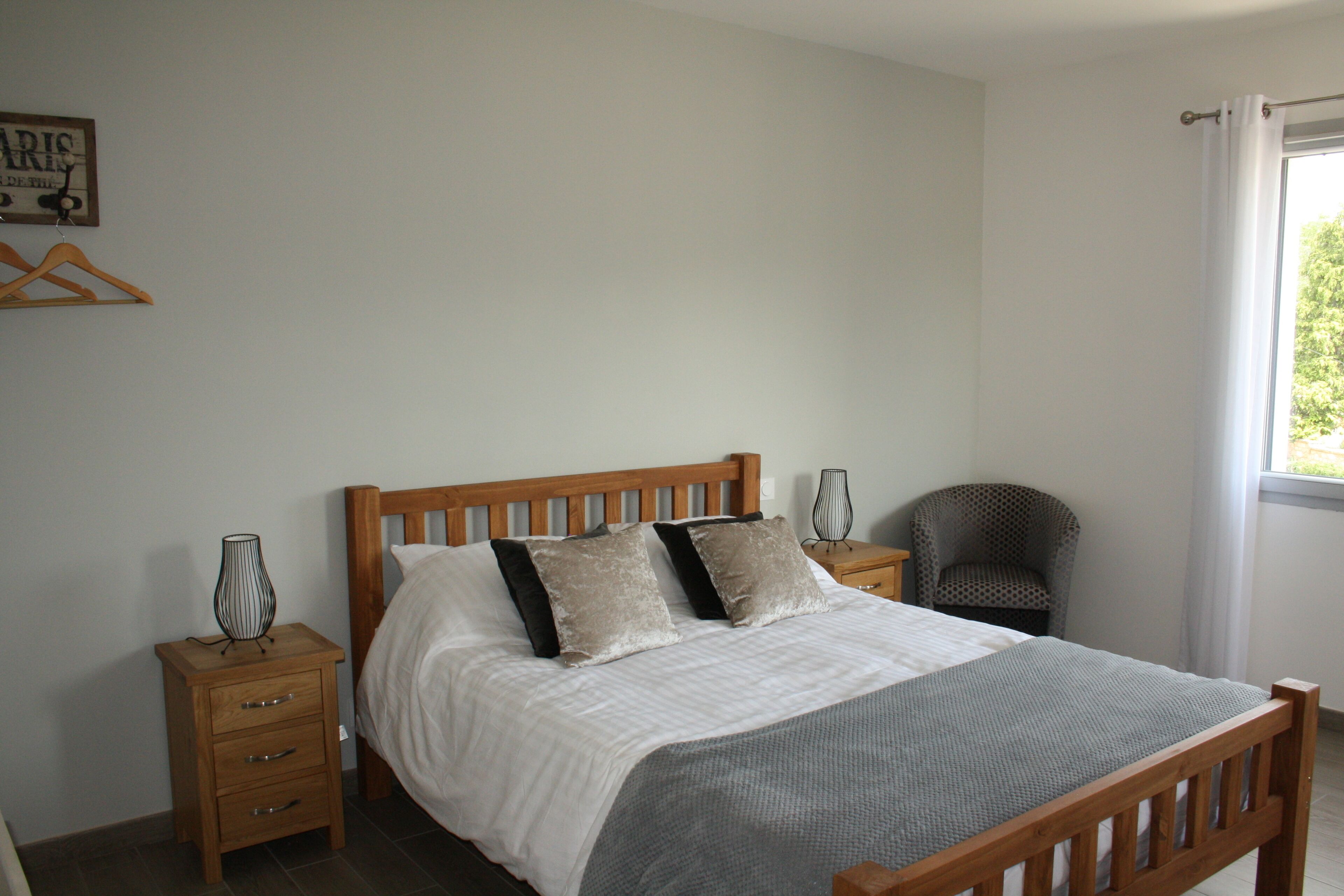 A bedroom with a wooden bed, a nightstand with drawers, a chair, and a window with curtains.