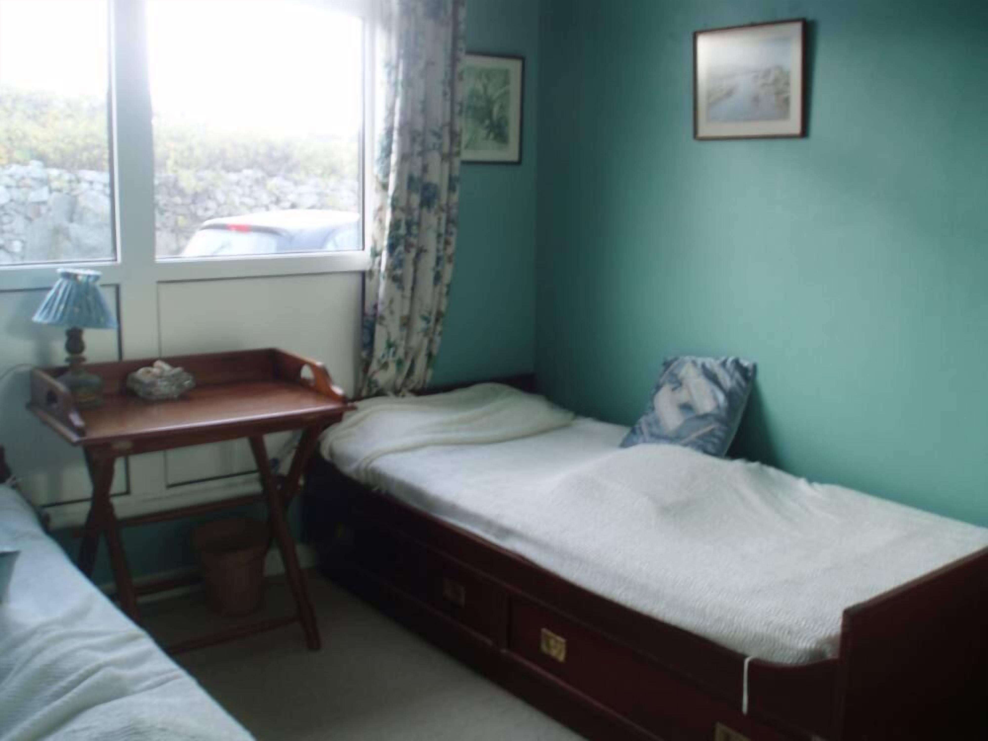 A small, single-bed room with a wooden desk and chair, a window with curtains, and a wall-mounted picture.