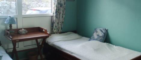 A small, single-bed room with a wooden desk and chair, a window with curtains, and a wall-mounted picture.