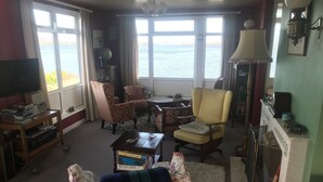 A living room with a view of the sea, a television, a bookshelf, and a lamp.