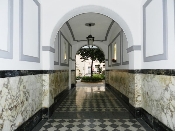 Um corredor com piso xadrez, paredes brancas com detalhes em mármore e teto em arco.