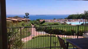 A balcony with a view of a swimming pool, a grassy area, and a distant body of water. - Stunning three-room lake view in a residence with three swimming pools and 36,000 m2 of greenery (Manerba del Garda)