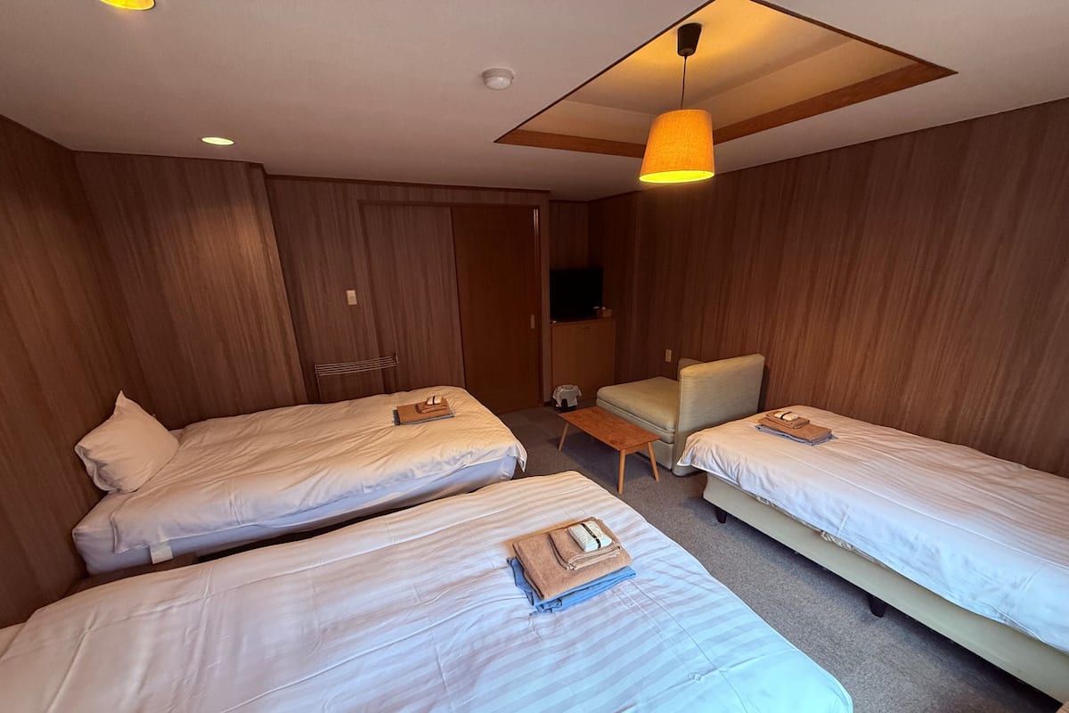 A hotel room with two single beds, a small table, a chair, and a ceiling light fixture.