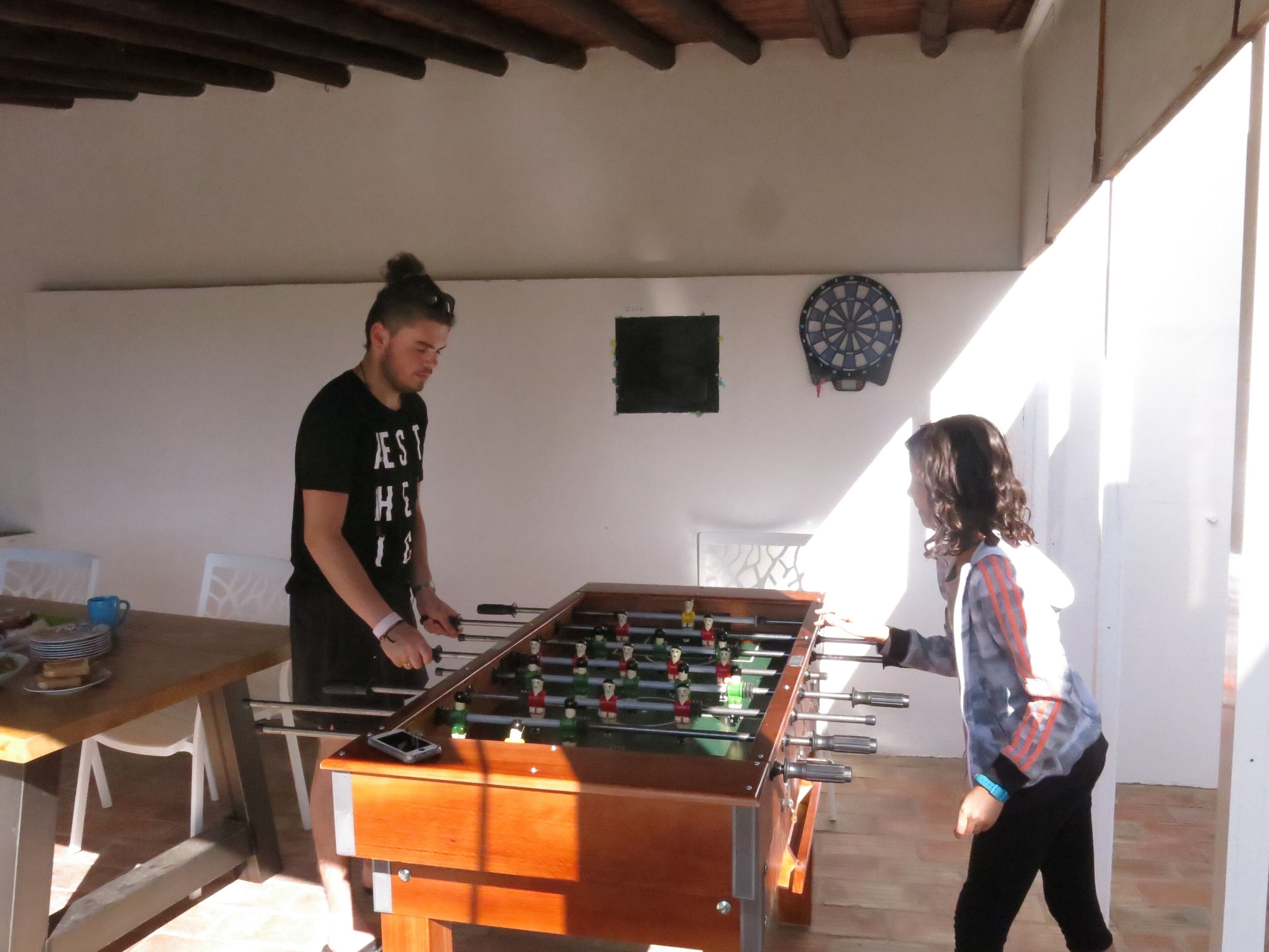 A foosball table in a room with a dartboard and a small table with items on it.