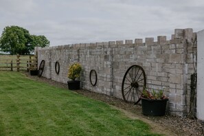 Un muro de piedra con aberturas circulares, plantas en macetas y una valla de madera en una zona cubierta de césped.