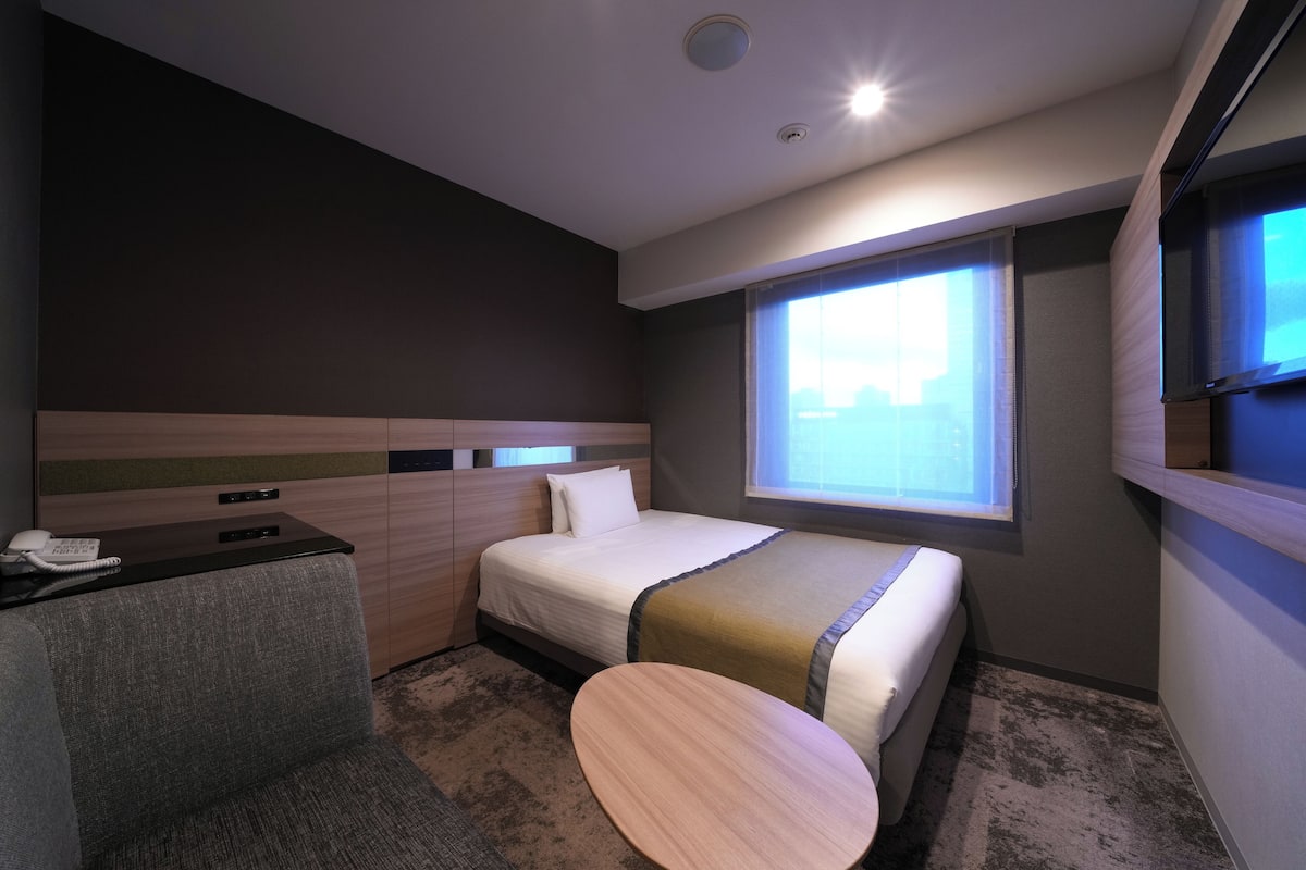 A hotel room with a bed, a desk with a telephone, a small round table, and a window with blinds.