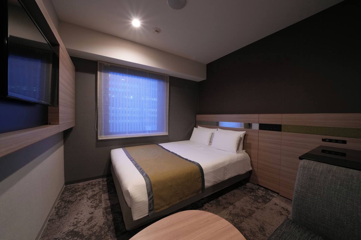 A hotel room with a bed, a small desk, a window with blinds, and a television on the wall.