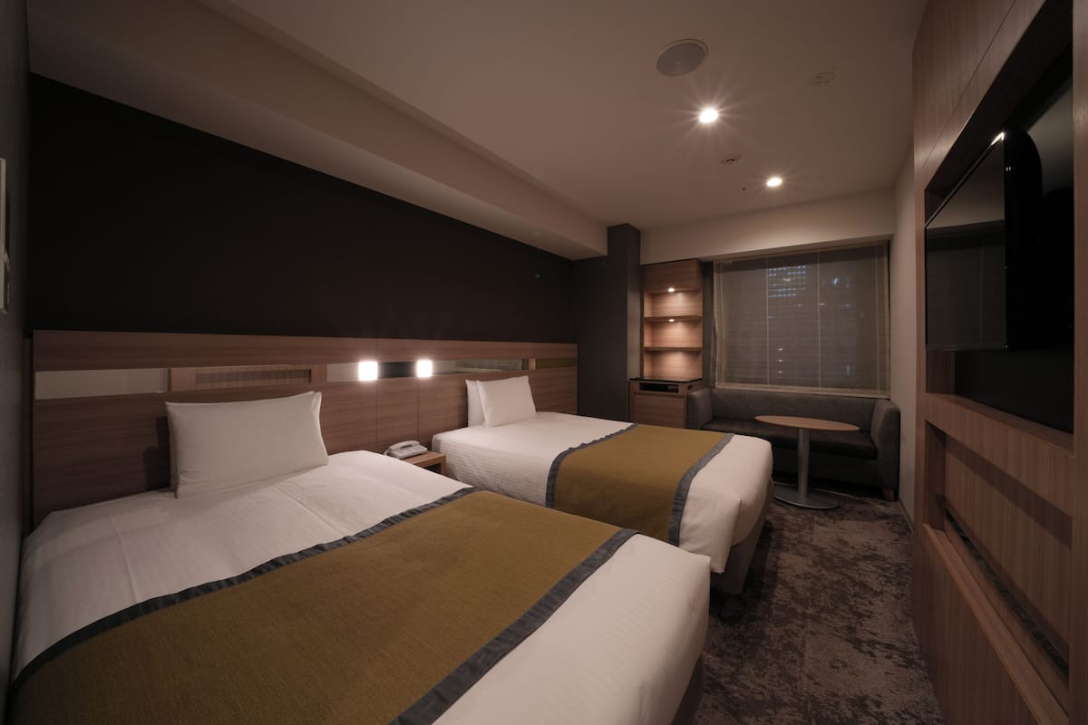 A hotel room with two beds, a small table, a TV, and a window with a city view.