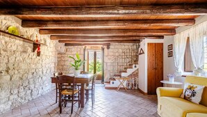 A rustic interior with a stone wall, wooden beams, a dining table with chairs, a yellow sofa, and a staircase leading to an upper level.