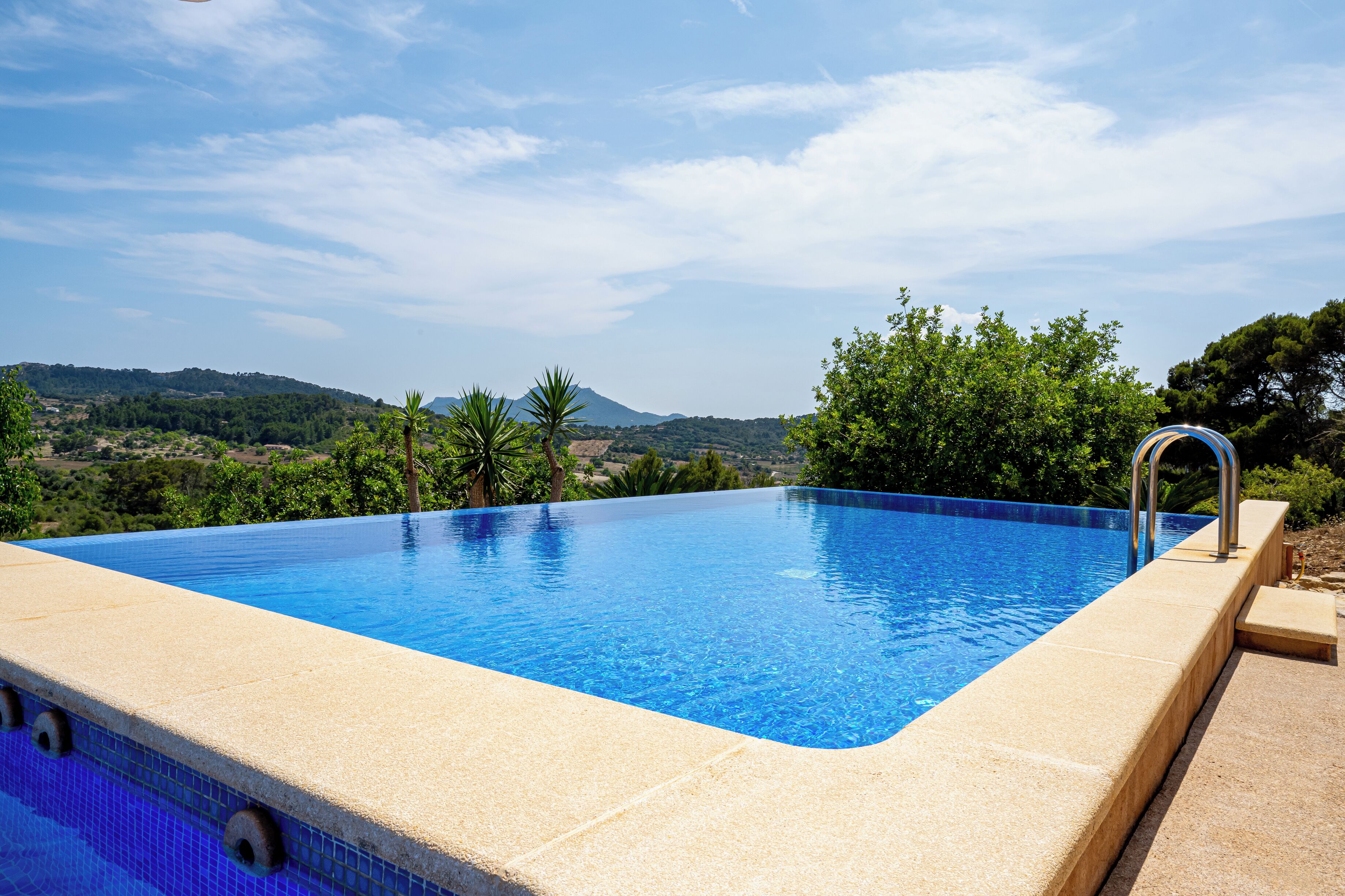 A rectangular infinity pool with a clear blue edge, surrounded by a beige deck, set against a backdrop of lush greenery and distant hills.