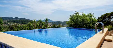A rectangular infinity pool with a clear blue edge, surrounded by a beige deck, set against a backdrop of lush greenery and distant hills.