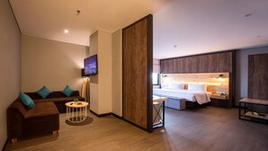Ein modernes Hotelzimmer mit einer Couch, einem kleinen runden Tisch, einem Bett mit Holz-Kopfstück, einem Fernseher und einem Nachttisch mit Lampe.