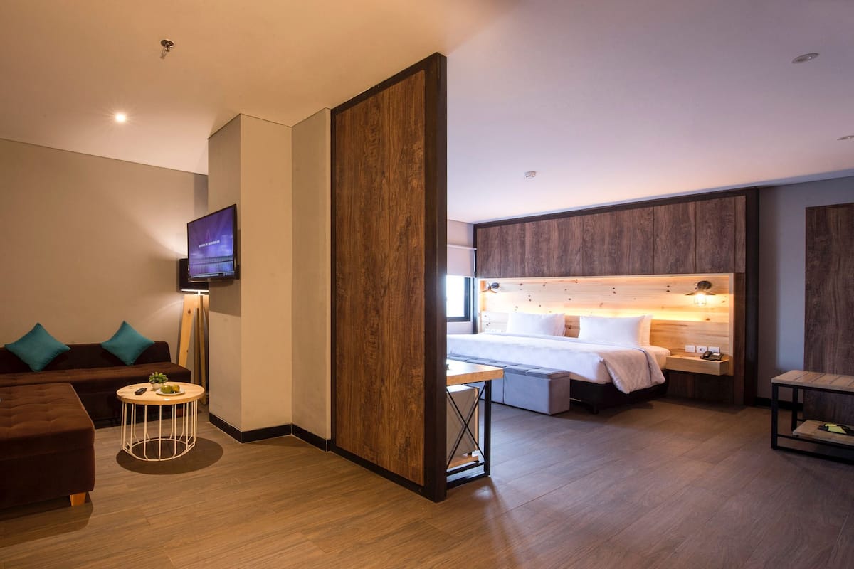 Una habitación de hotel moderna con un sofá, una mesita redonda pequeña, una cama con cabecero de madera, un televisor y una mesita de noche con una lámpara.