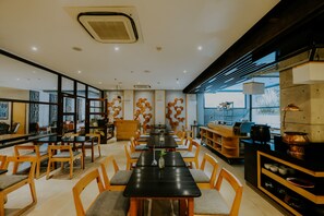 Ein modernes Restaurant mit langen Tischen und Holzstühlen, einem Küchenbereich mit Kupfergeschirr und großen Fenstern, die für natürliches Licht sorgen.