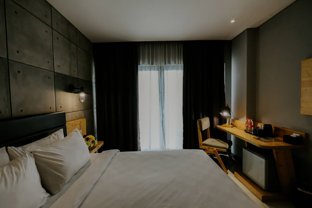 Habitación de hotel con una cama grande, un escritorio con una silla, una mesita de noche con lámpara y una ventana con cortinas.