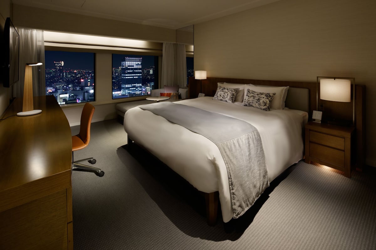 Habitación de hotel con una cama grande, un escritorio, una silla y vista a la ciudad de noche.