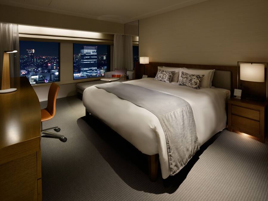 A hotel room with a large bed, a desk, a chair, and a view of the city at night.