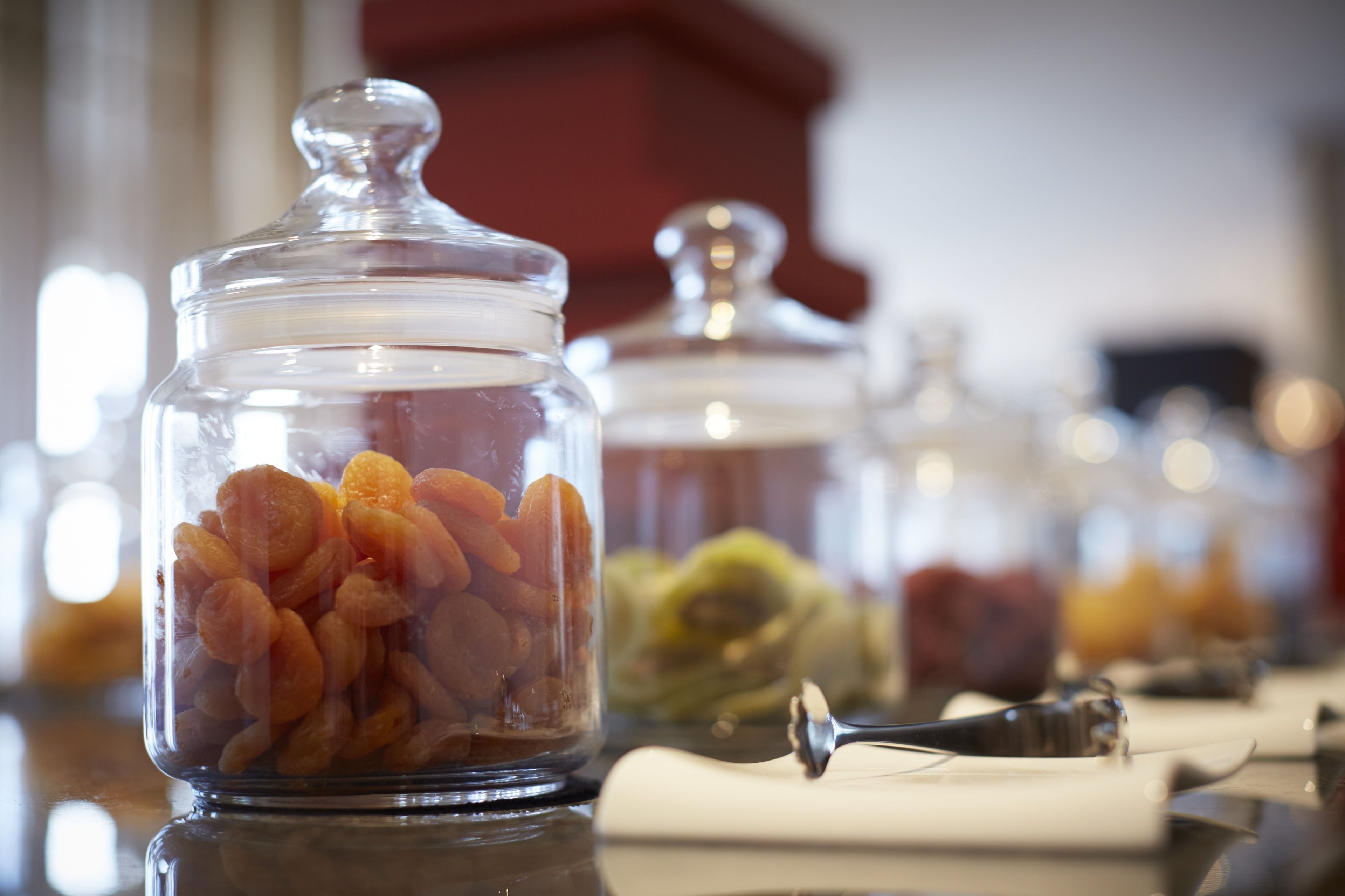 A glass jar filled with dried apricots on a table with a spoon and a napkin.