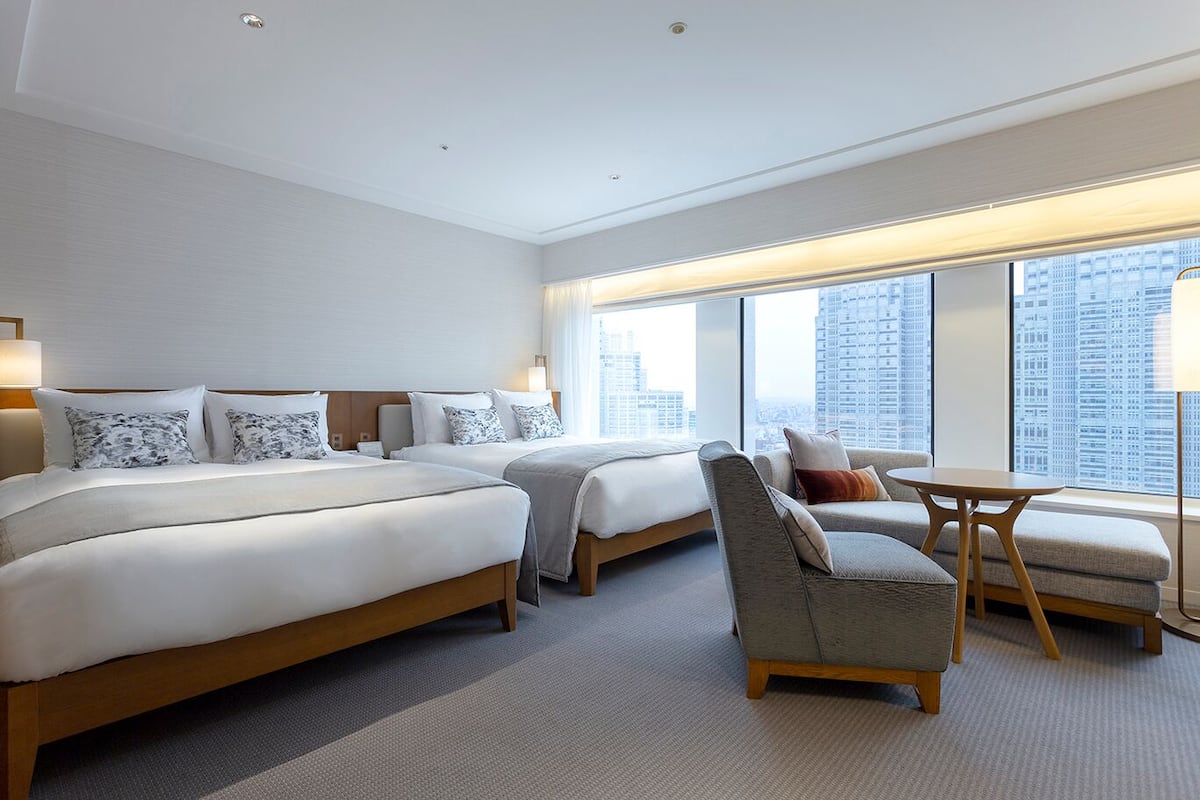 Habitación de hotel con dos camas, un sofá, una mesa y grandes ventanales con vista a la ciudad.