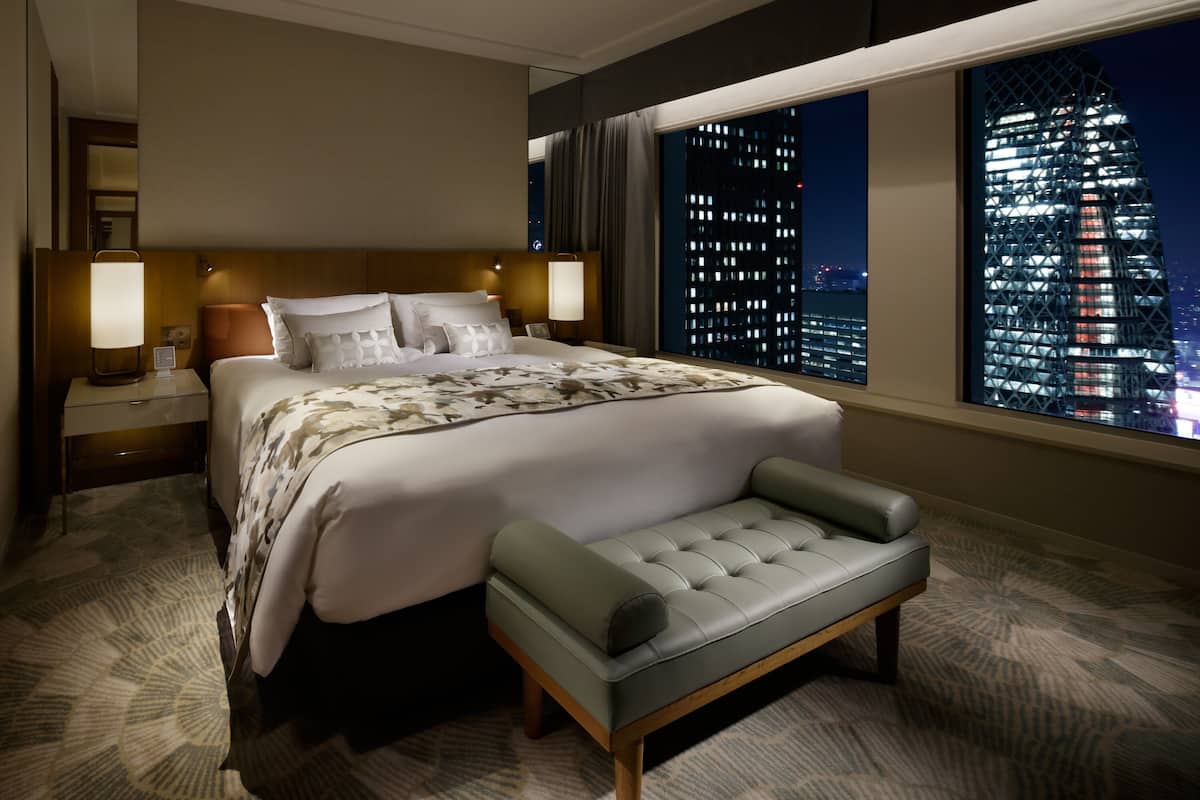 Habitación de hotel con una cama grande, un banco, una mesita de noche, una lámpara y vistas a la ciudad de noche.