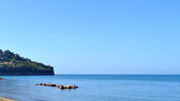 Un paysage côtier avec une rive rocheuse, une mer calme et une colline parsemée de bâtiments.