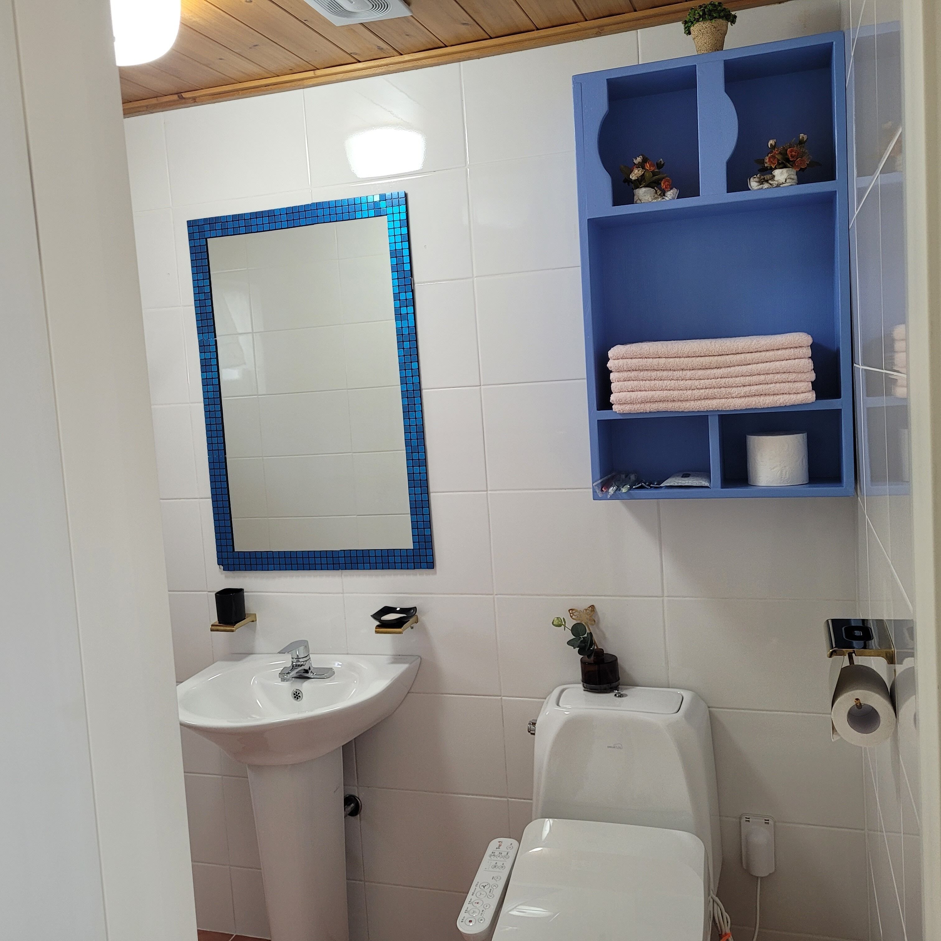 Casa de banho com lavatório branco, sanita, espelho com moldura azul e prateleira azul com toalhas e produtos de higiene pessoal.