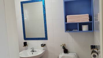 Casa de banho com lavatório branco, sanita, espelho com moldura azul e prateleira azul com toalhas e produtos de higiene pessoal.