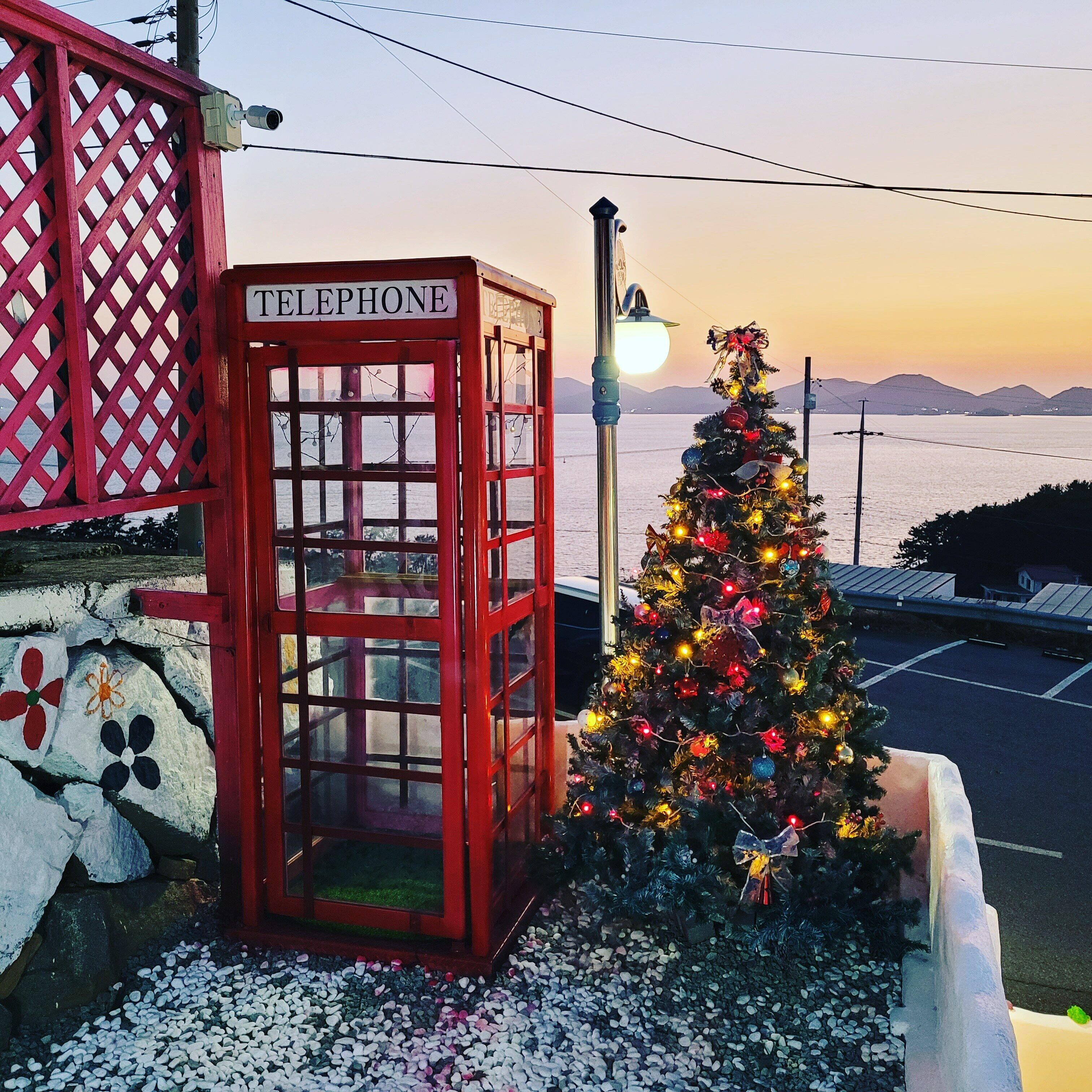 A red telephone booth with a sign, a decorated Christmas tree, and a view of the sea.
