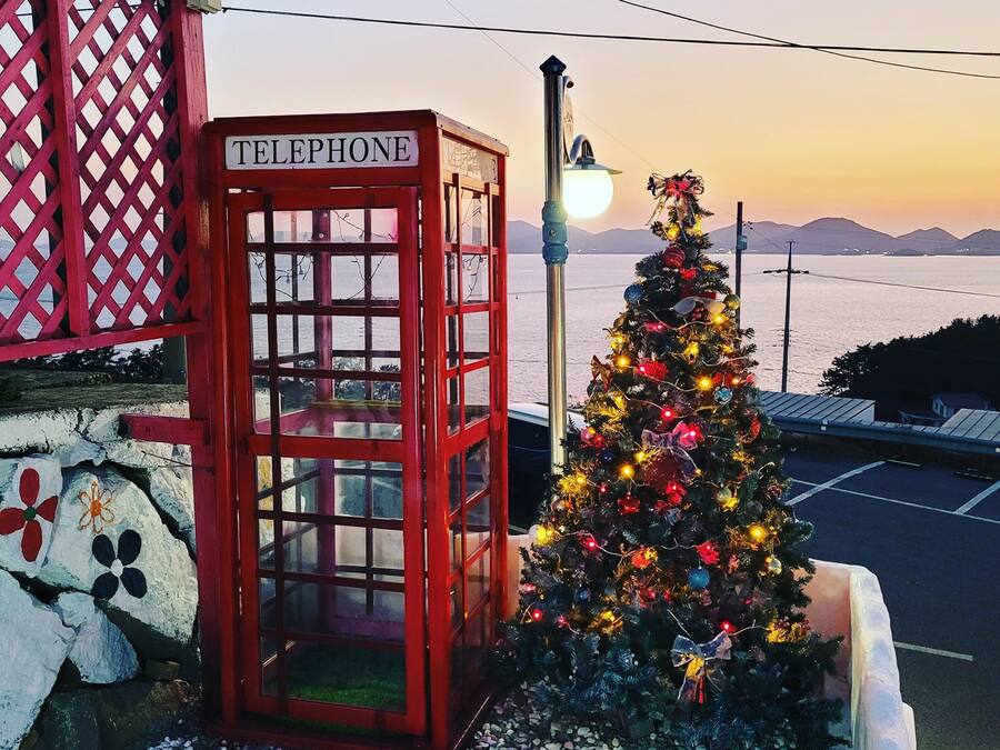 A red telephone booth with a sign, a decorated Christmas tree, and a view of the sea.