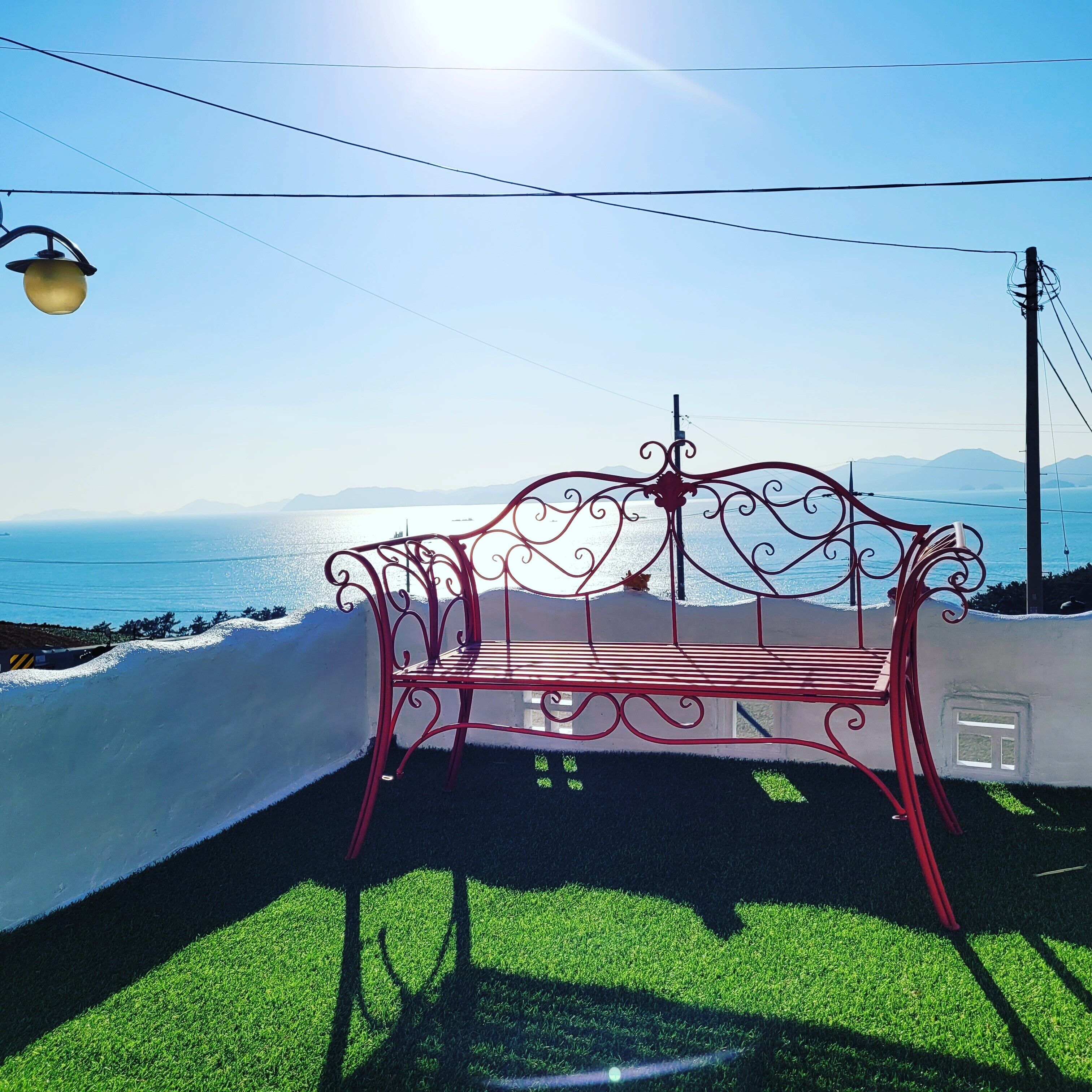 A red metal bench with a white cushion, positioned on a grassy area with a view of the sea and mountains in the background.