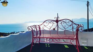 A red metal bench with a white cushion, positioned on a grassy area with a view of the sea and mountains in the background.