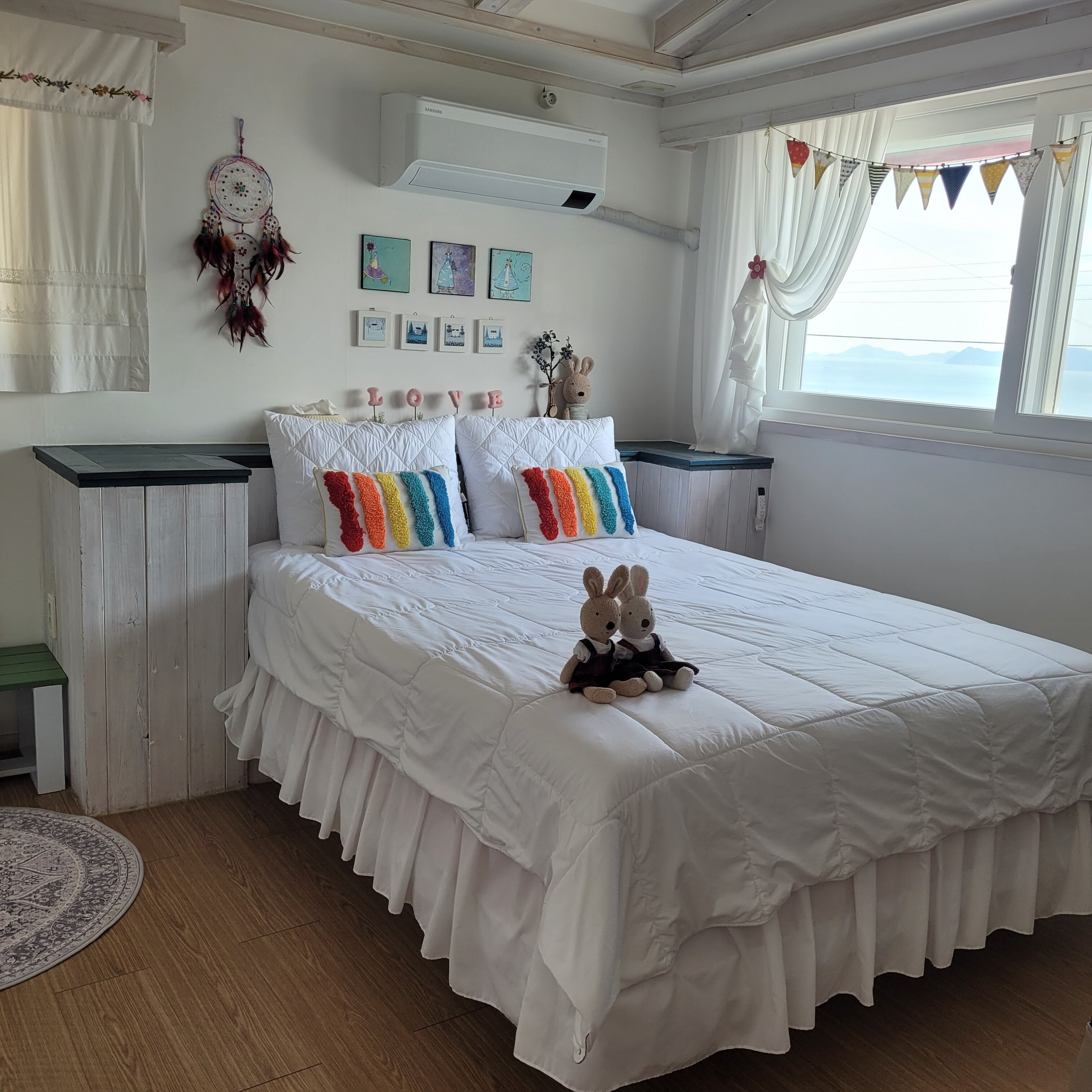 A bedroom with a large bed, white bedding, a dreamcatcher, and a view of the sea through a window.