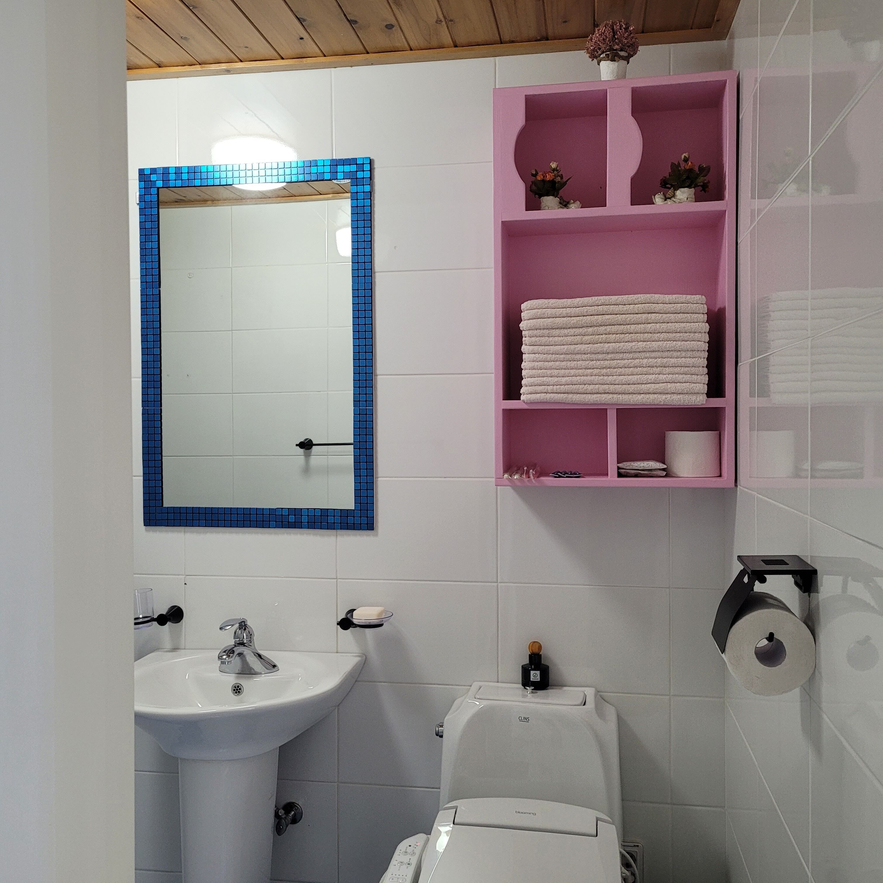 Een badkamer met een wit wastafelmeubel, een toilet, een spiegel met blauwe lijst en een roze plank met handdoeken.