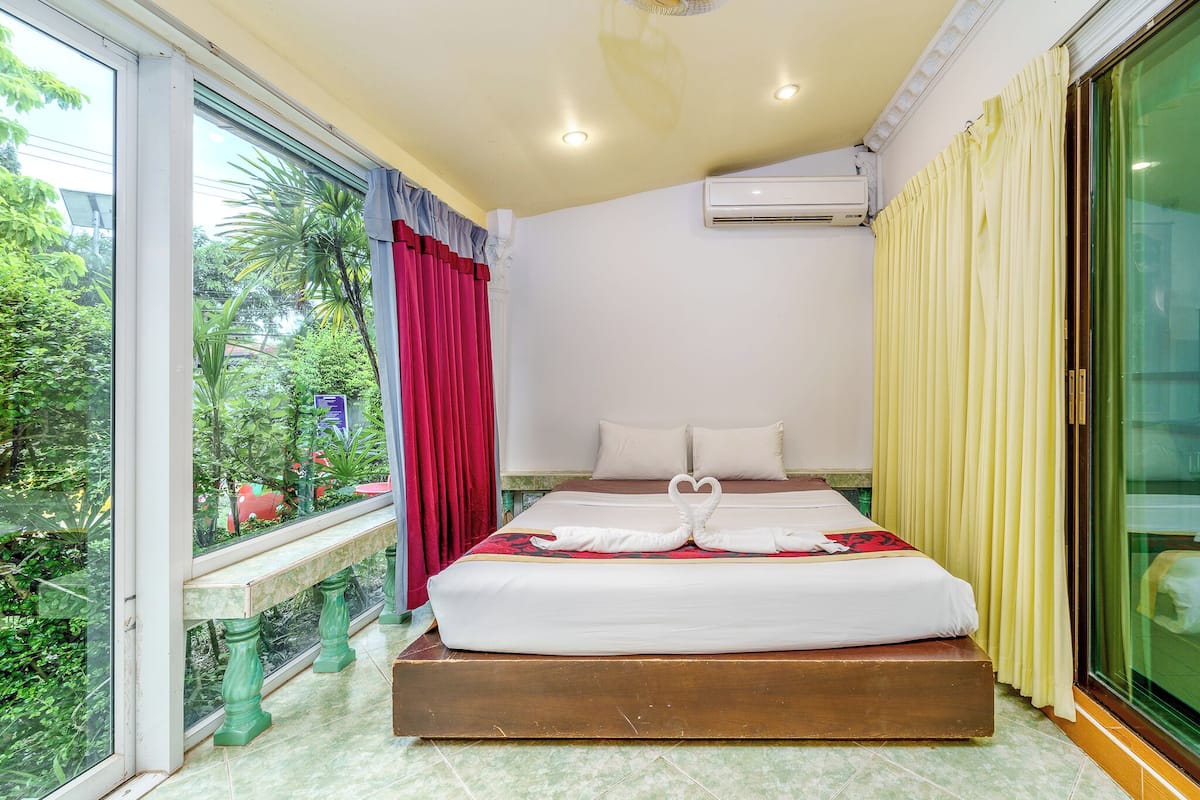 A hotel room with a bed, a window with curtains, an air conditioning unit, and a view of greenery outside.