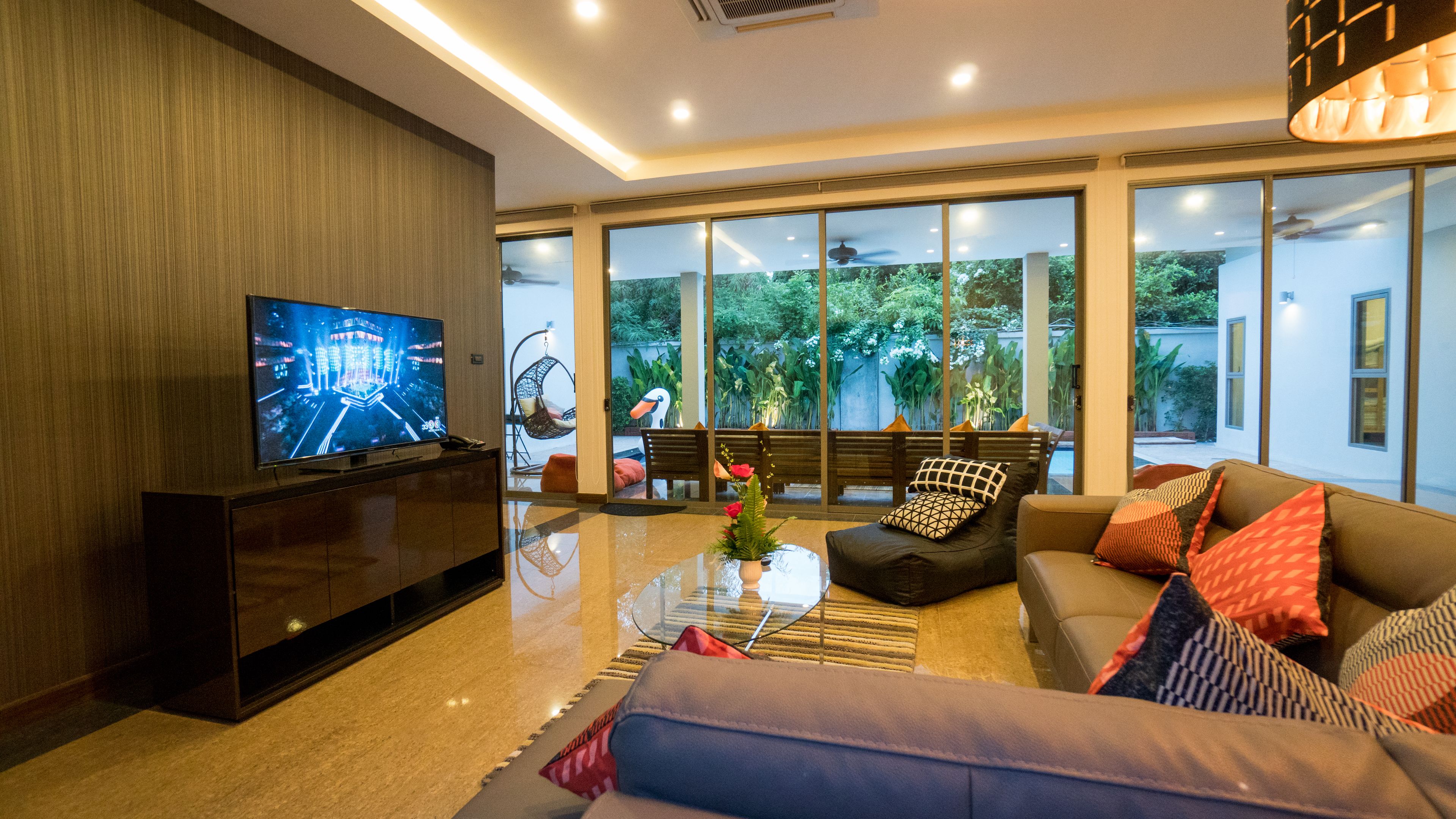 Ein modernes Wohnzimmer mit Fernseher, bequemen Sitzmöglichkeiten, einem Glastisch und großen Fenstern mit Blick auf Grünflächen.