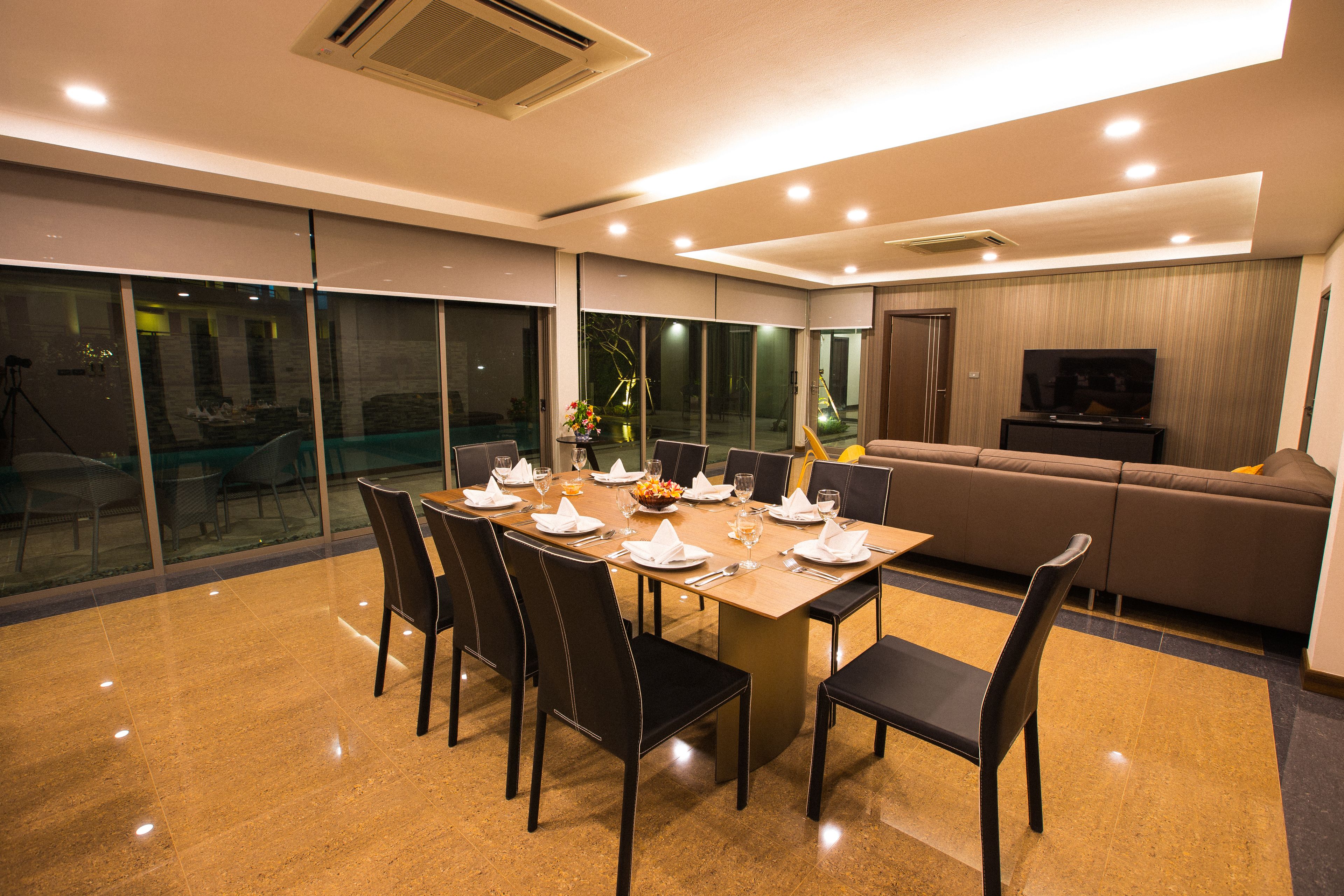 A modern dining area with a large table set for six, surrounded by chairs, and a sofa in the background.