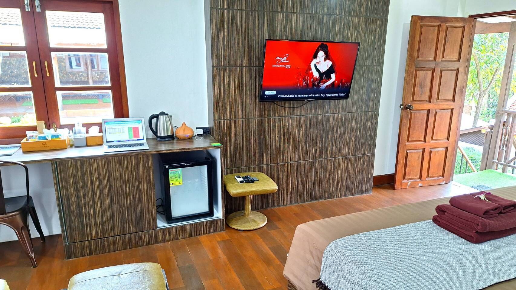 Habitación de hotel con escritorio de madera, televisor montado en la pared, refrigerador pequeño, taburete acolchado y cama con manta.