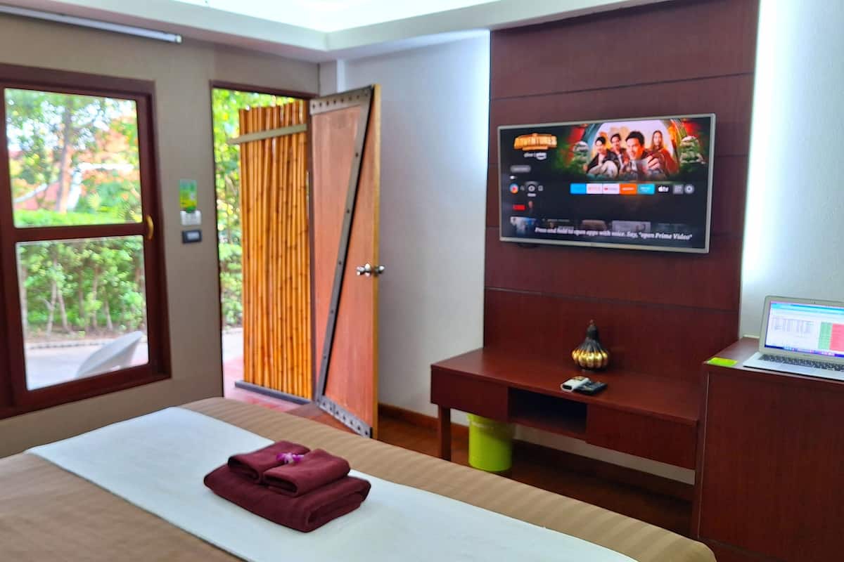 A hotel room with a bed, a TV on the wall, and a window with a view of greenery.