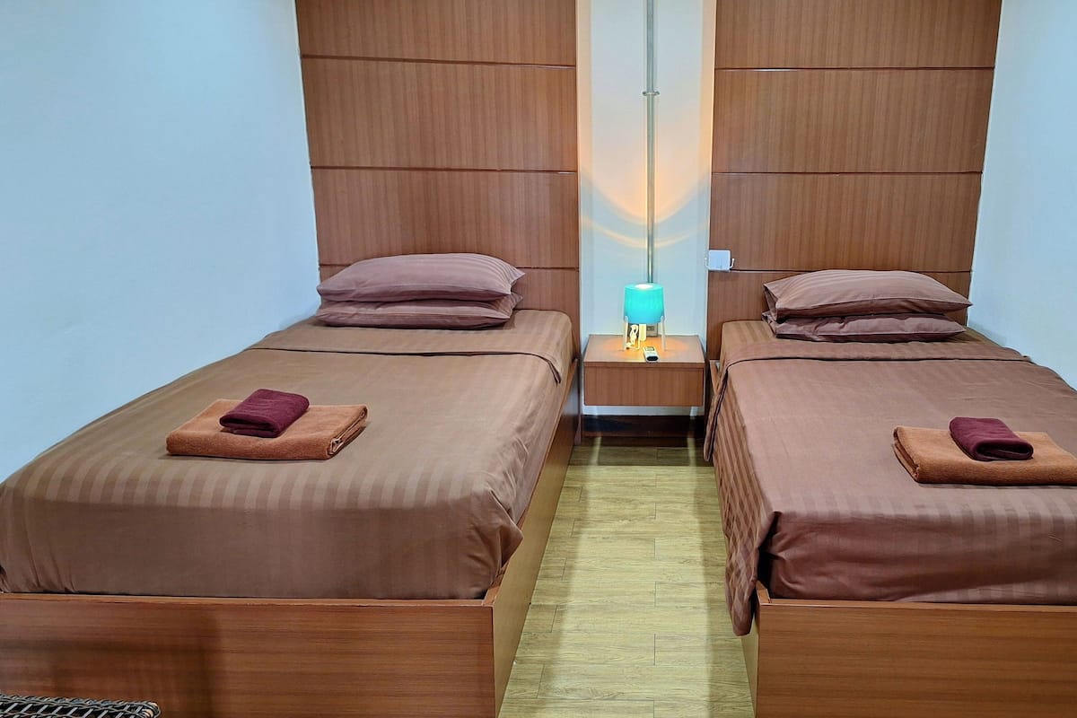 A room with two beds, each with a pillow and a folded towel. There is a nightstand with a lamp between the beds.